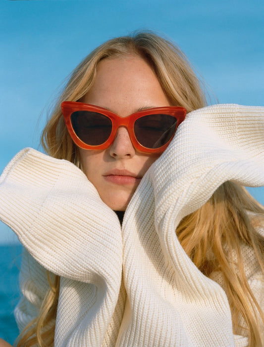 Woman wearing stylish red acetate sunglasses and a white sweater, with a serene blue sky in the background.